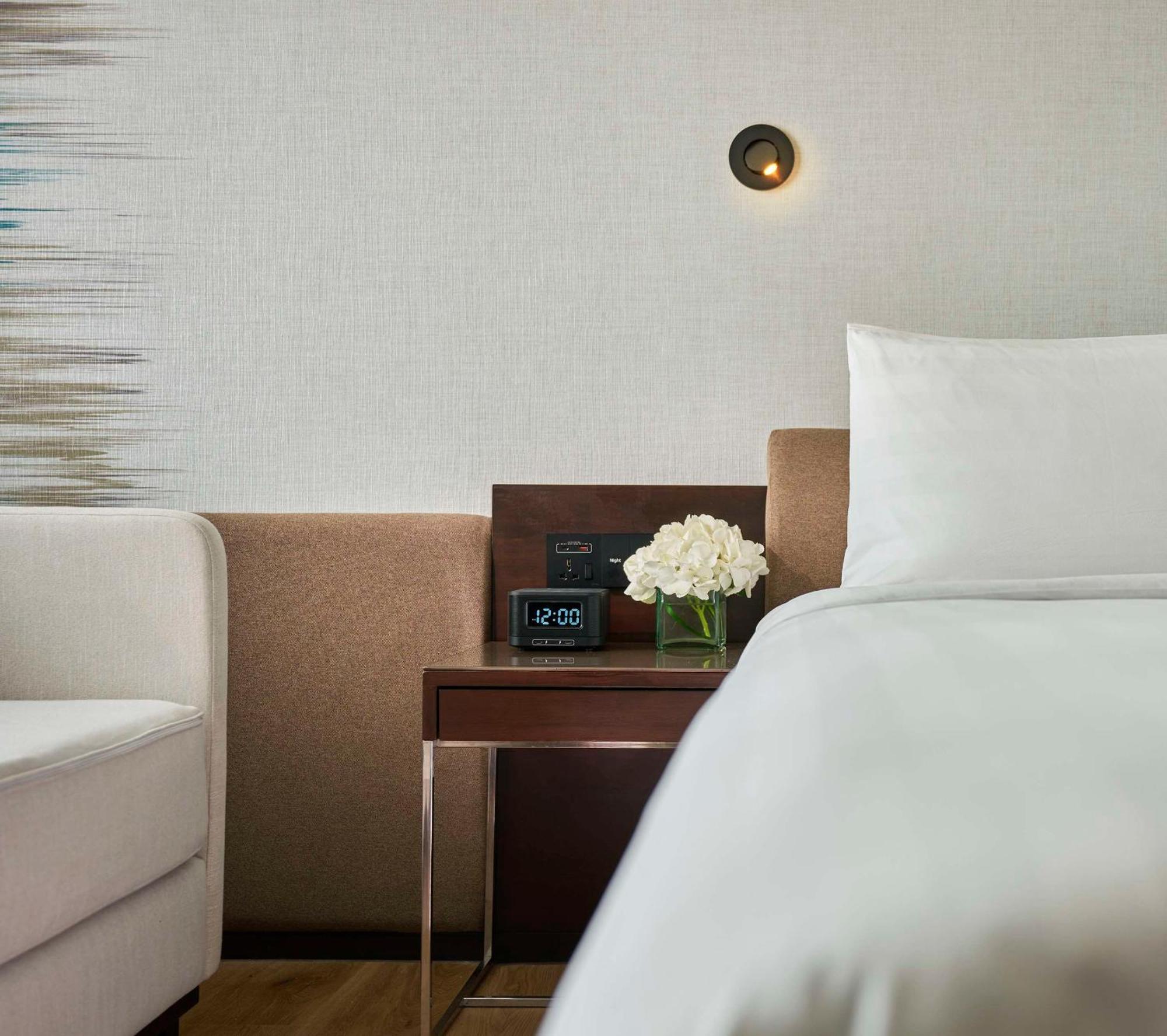 Traders Hotel, Κουάλα Λουμπούρ Εξωτερικό φωτογραφία The photo shows a cozy hotel room corner featuring a bed with white bedding. Next to the bed is a small wooden nightstand that has a digital clock displaying the time. On the nightstand, there is a small bouquet of white flowers, and beside it, you c