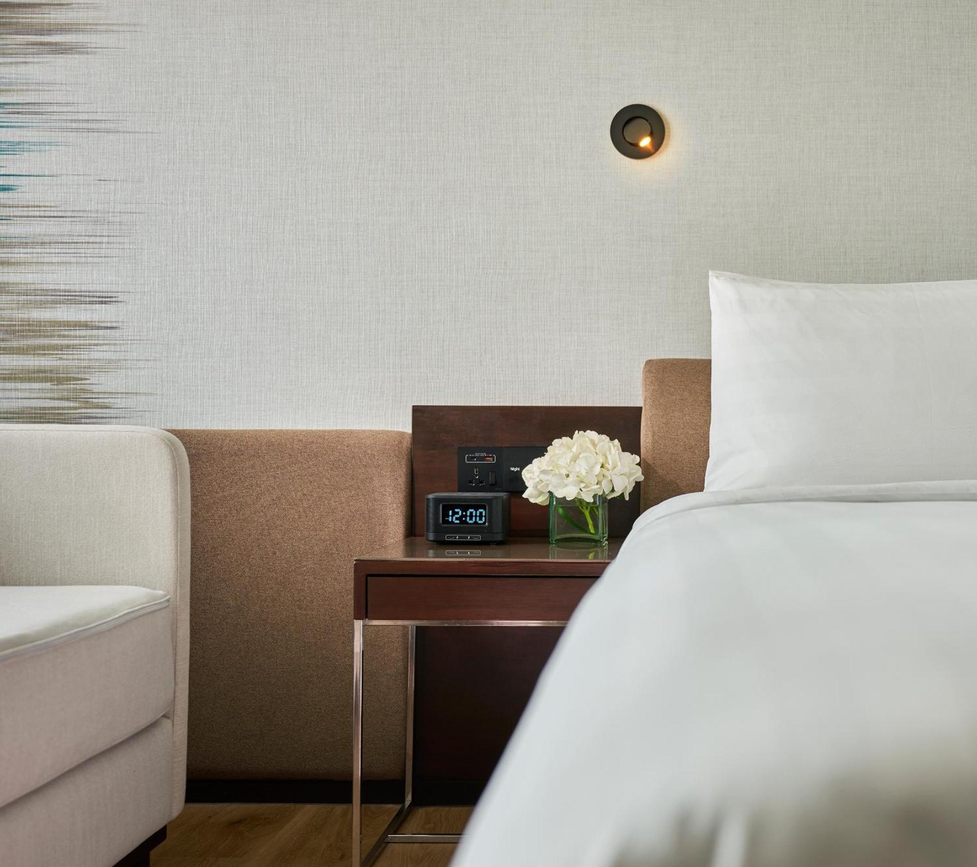 Traders Hotel, Κουάλα Λουμπούρ Εξωτερικό φωτογραφία The photo shows a cozy bedroom setting. To the right, there's a neatly made bed with white bedding. Next to the bed, there's a wooden bedside table with a small vase of white flowers on it. On the table, there is also a digital clock displaying the t